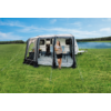 Witte caravan met een opgezet voortent op een grasveld naast een meer; een persoon staat in de open ingang van de voortent en houdt een beker vast; een stoel en tafel zijn zichtbaar in de voortent; blauwe lucht met wolken en een rij bomen aan de horizon