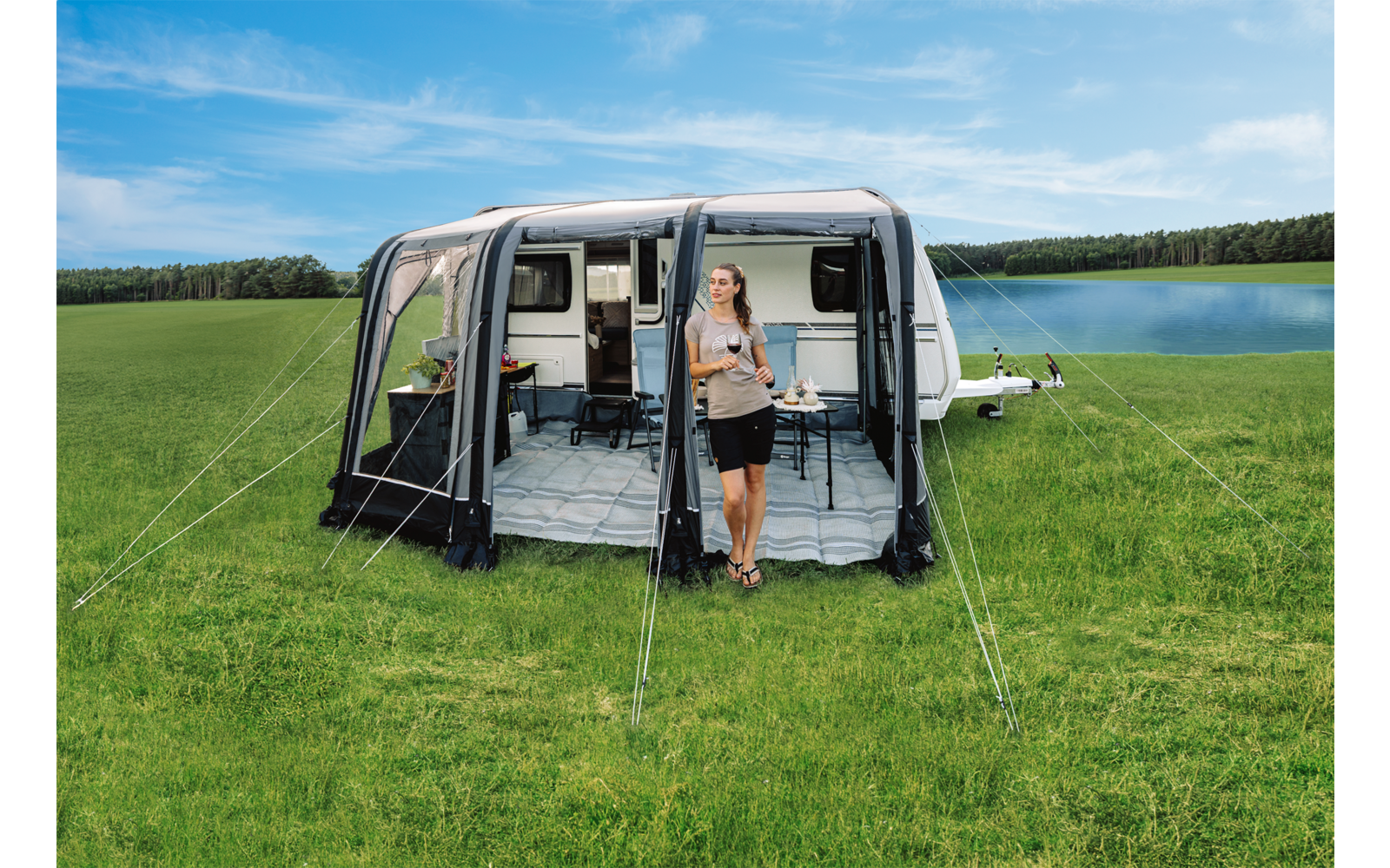 Witte caravan met een opgezet voortent op een grasveld naast een meer; een persoon staat in de open ingang van de voortent en houdt een beker vast; een stoel en tafel zijn zichtbaar in de voortent; blauwe lucht met wolken en een rij bomen aan de horizon