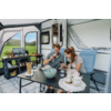 Twee volwassenen (een man en een vrouw) zitten aan een zwarte tafel voor een witte caravan en houden glazen vast. Op de tafel: een kom, een fles, broodstengels, borden en kopjes; deur van de caravan open; groen veld op de achtergrond