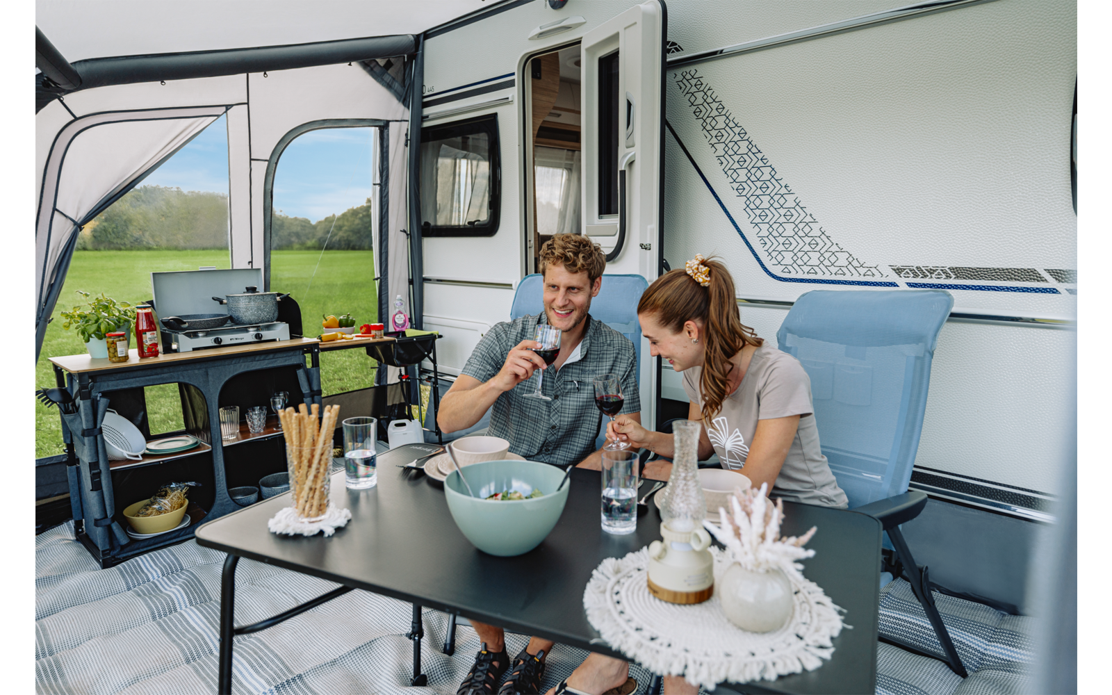 Twee volwassenen (een man en een vrouw) zitten aan een zwarte tafel voor een witte caravan en houden glazen vast. Op de tafel: een kom, een fles, broodstengels, borden en kopjes; deur van de caravan open; groen veld op de achtergrond