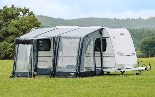 White caravan with a gray awning attached, on a green field with trees and hills in the background