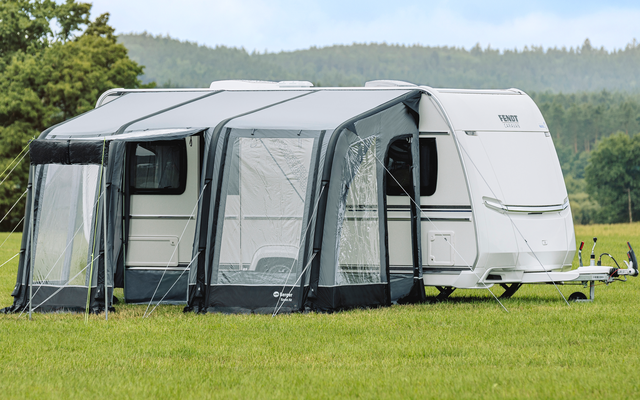 Caravane blanche avec un auvent gris posé sur un champ vert, arbres et collines en arrière-plan