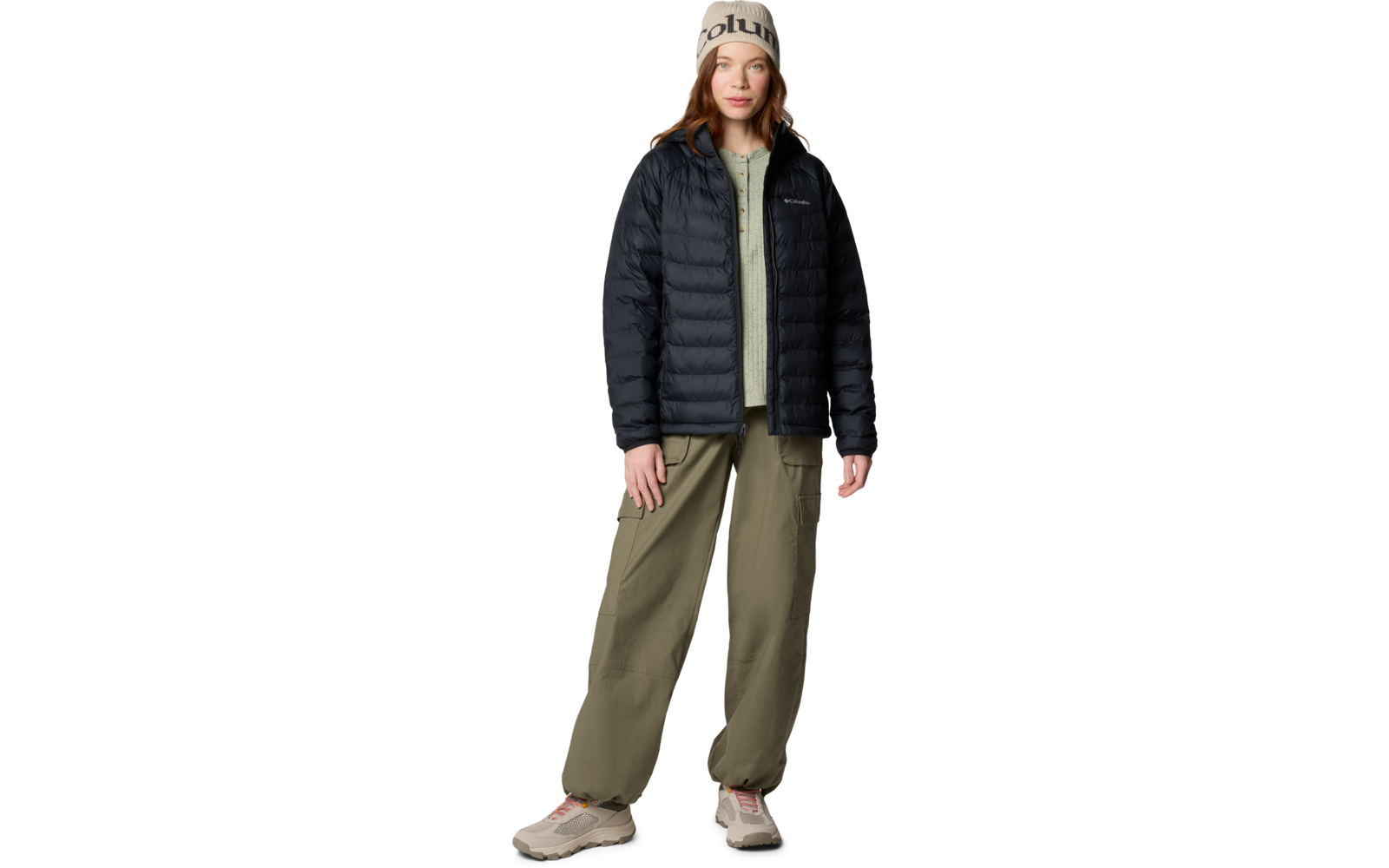 Personne debout de face portant un bonnet beige avec inscription, une doudoune noire matelassée, un pull zippé vert clair, un pantalon cargo vert olive et des baskets beiges; cheveux bruns mi-longs; fond blanc
