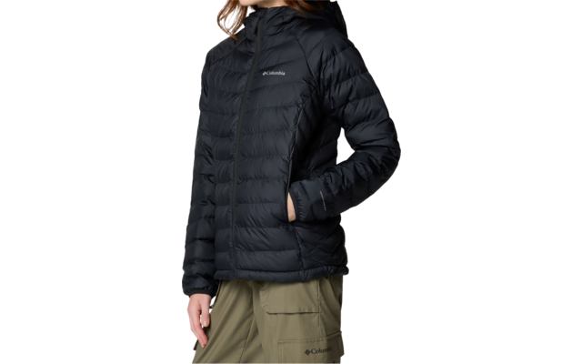 Personne aux cheveux bruns mi-longs portant une doudoune noire matelassée à capuche et fermeture éclair, mains dans les poches, pantalon vert olive, fond blanc