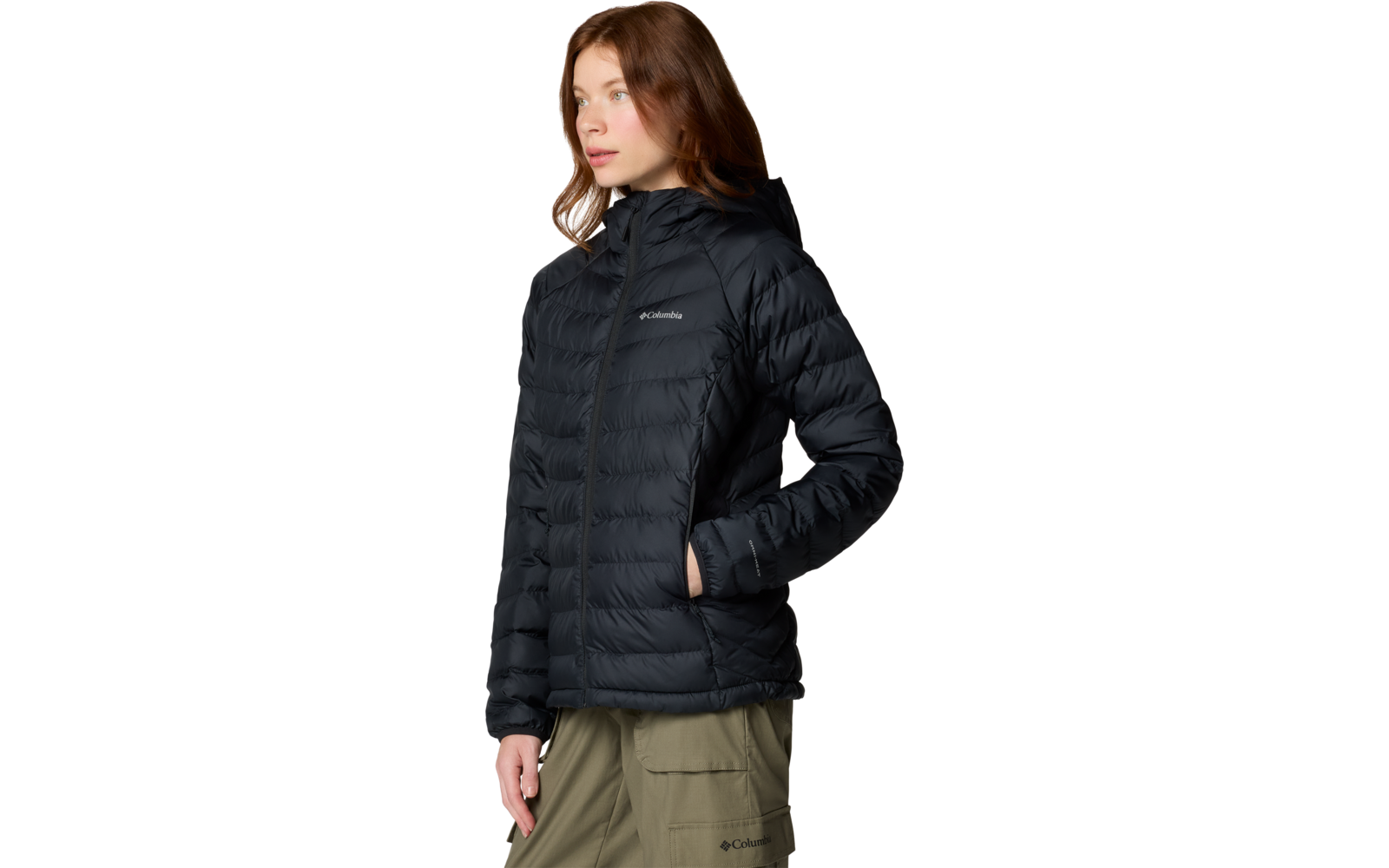 Personne aux cheveux bruns mi-longs portant une doudoune noire matelassée à capuche et fermeture éclair, mains dans les poches, pantalon vert olive, fond blanc