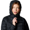Personne portant une doudoune noire avec capuche relevée, main droite tenant la fermeture éclair près du menton, regard légèrement vers la gauche, logo Columbia visible sur la poitrine, fond blanc