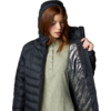 Femme aux cheveux roux portant une doudoune noire à capuche, tenant la veste ouverte ; pull vert en maille avec patte de boutons visible