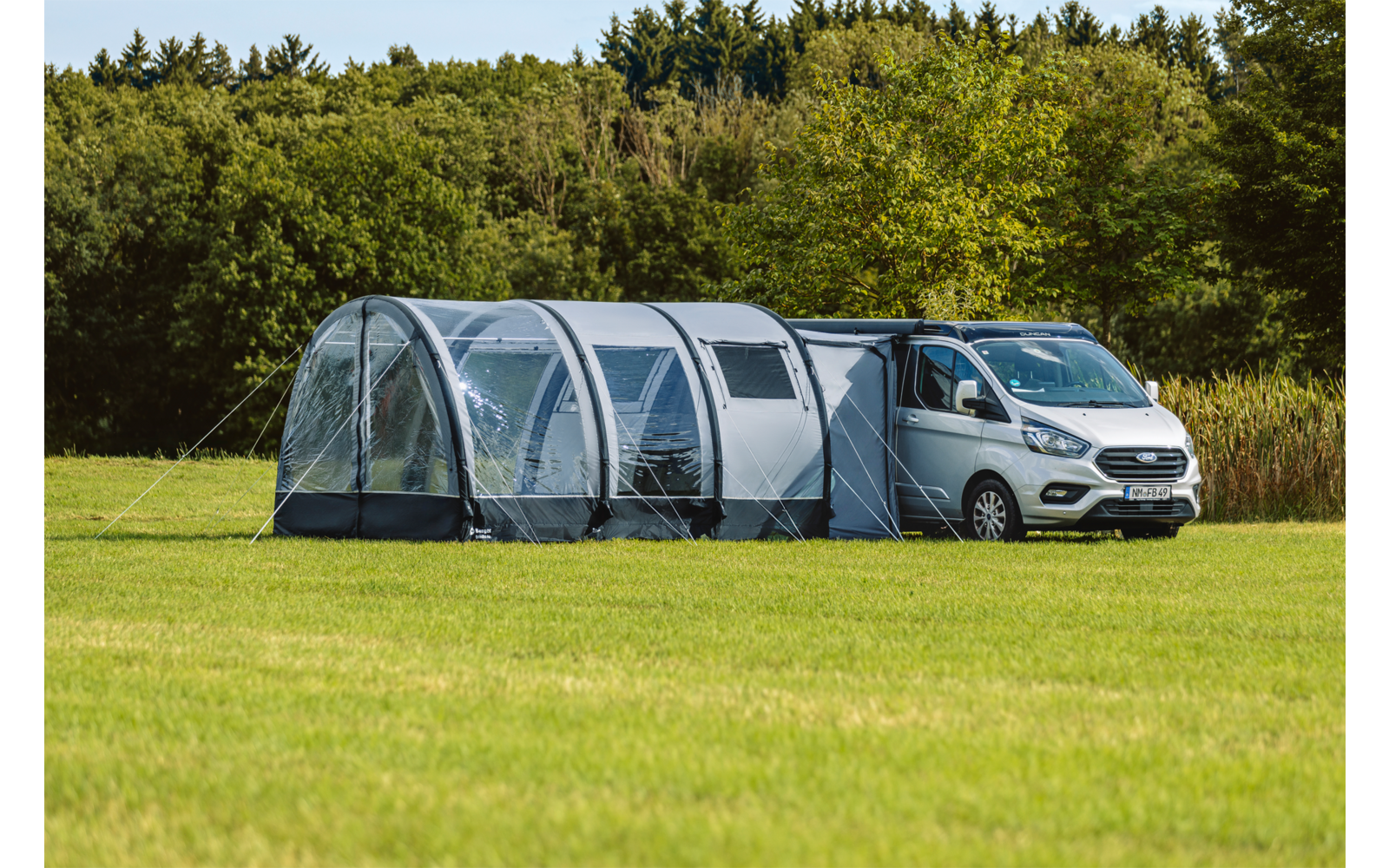 Weißer Van neben großem grauem Tunnelzelt auf einer Wiese, im Hintergrund Bäume