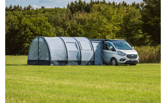 Weißer Transporter mit angebautem grauem Tunnelvorzelt auf einer grünen Wiese, Bäume im Hintergrund