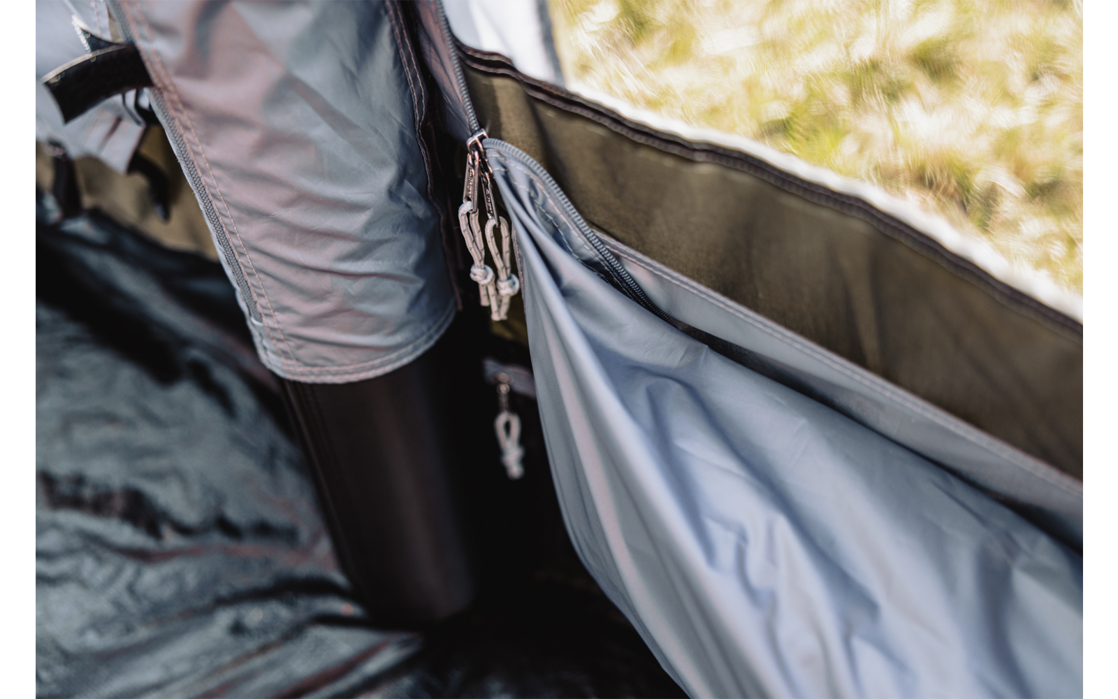 Geöffnetes Zelt mit offenem Reißverschluss, an dem mehrere Schlüssel hängen; graues Innenzeltmaterial; links ein schwarzer Trinkbecher; Gras im Hintergrund sichtbar