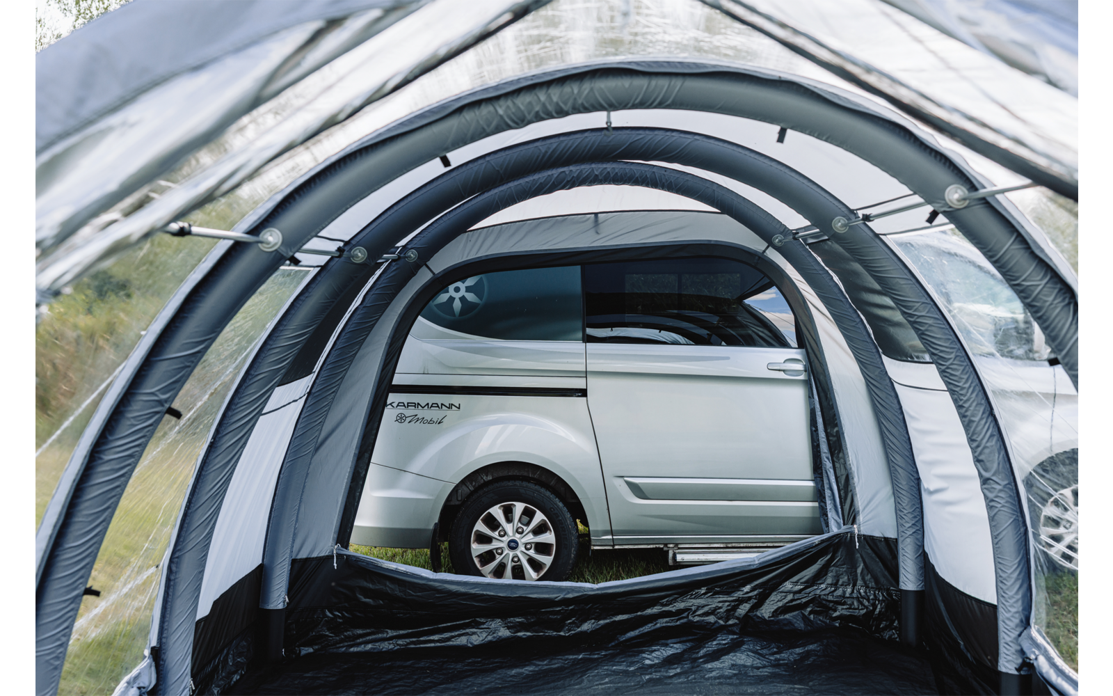 Blick aus dem Inneren eines aufblasbaren Vorzelts mit grauen Luftbögen und schwarzem Boden; draußen ein silberner Van mit sichtbarem Rad und Gras