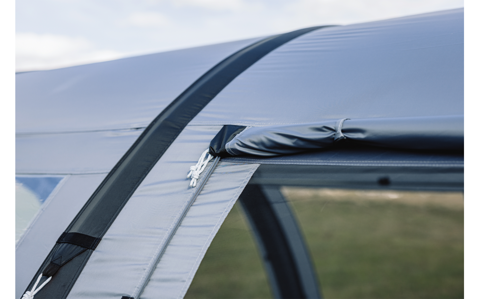 Nahaufnahme eines hellgrauen Stoffbereichs mit Reißverschluss, aufgerolltem Stoffriegel, Metall-Reißverschlussanhänger und schwarzem Verstärkungsstreifen; unscharfe Wiese und Himmel im Hintergrund