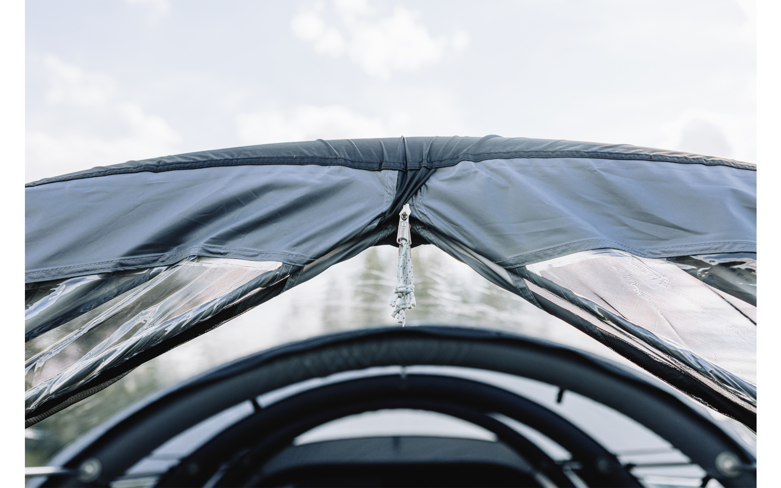 Geschlossene transparente Plastikplane mit Reißverschluss oben, zwei seitliche durchsichtige Fenster, gebogene schwarze Rahmenstangen im Vordergrund, heller wolkiger Himmel im Hintergrund