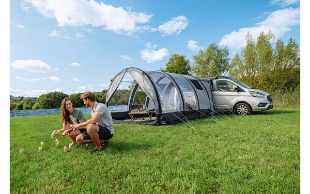 Zwei Personen hocken neben zwei kleinen Hunden auf einer Wiese; rechts ein silberner Van mit angebautem Tunnelzelt, See, Bäume und blauer Himmel mit Wolken
