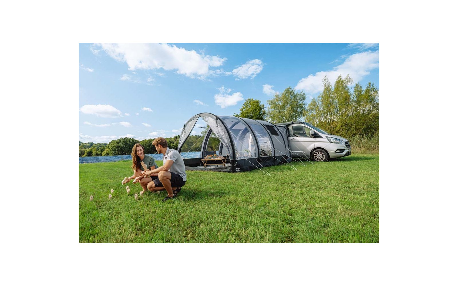 Zwei Personen hocken neben zwei kleinen Hunden auf einer Wiese; rechts ein silberner Van mit angebautem Tunnelzelt, See, Bäume und blauer Himmel mit Wolken