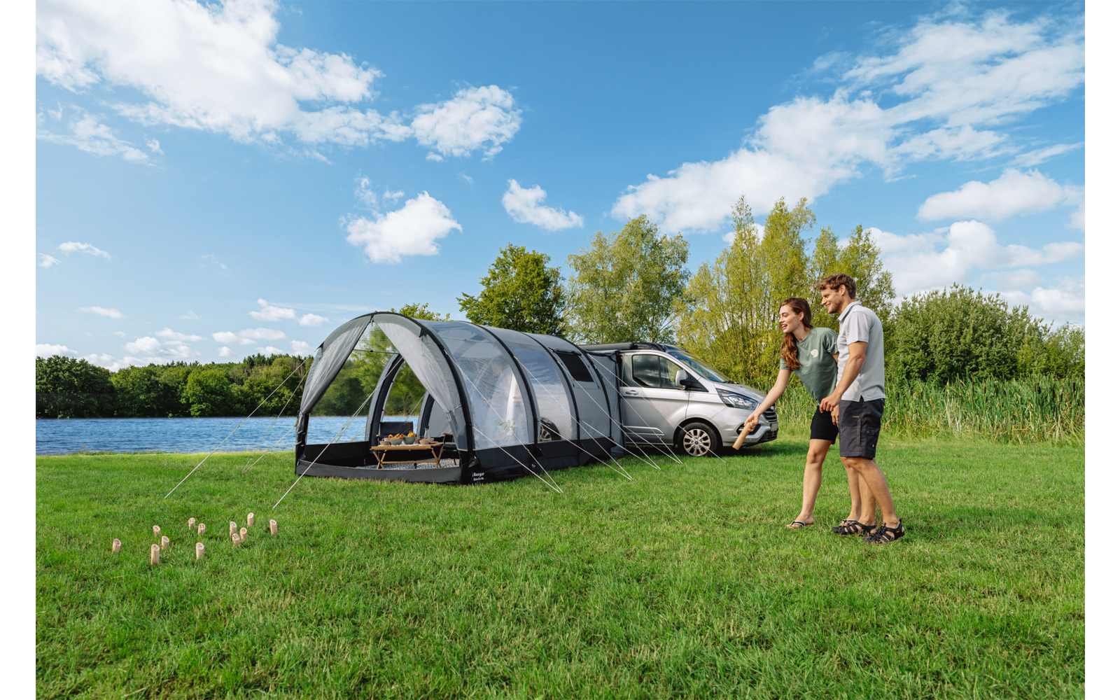 Two people walk on a grassy area beside a large pitched camping tent with a car next to it; lake, trees, and a blue sky with clouds in the background