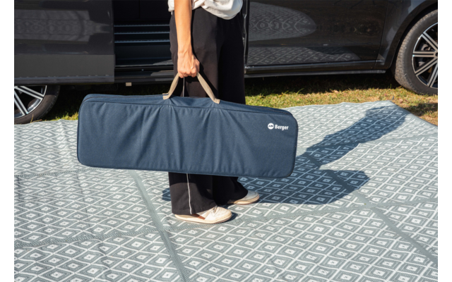 Metà inferiore del corpo di una persona che tiene per i manici una lunga borsa blu accanto a un'auto parcheggiata su un pavimento a motivo