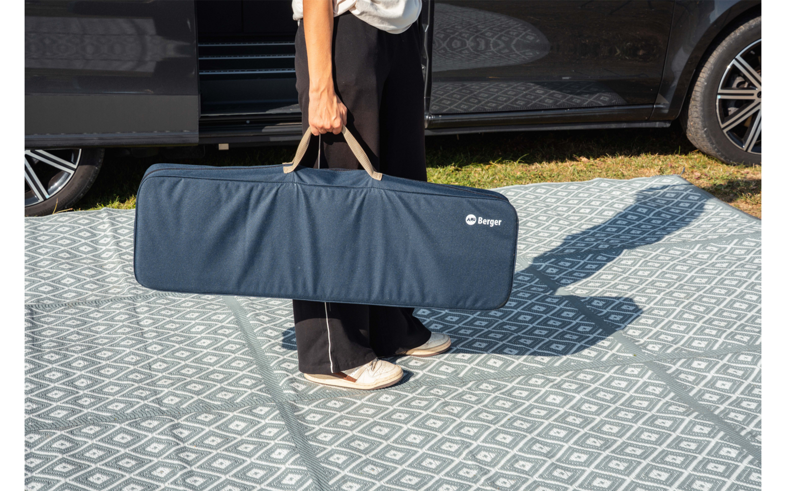 Metà inferiore del corpo di una persona che tiene per i manici una lunga borsa blu accanto a un'auto parcheggiata su un pavimento a motivo