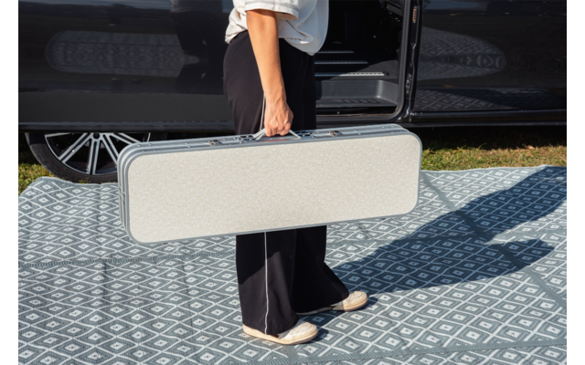 Parte inferiore del corpo di una persona con scarpe bianche e pantaloni neri che tiene una valigetta rettangolare grigio chiaro accanto a una portiera di auto nera aperta su un tappeto a motivi