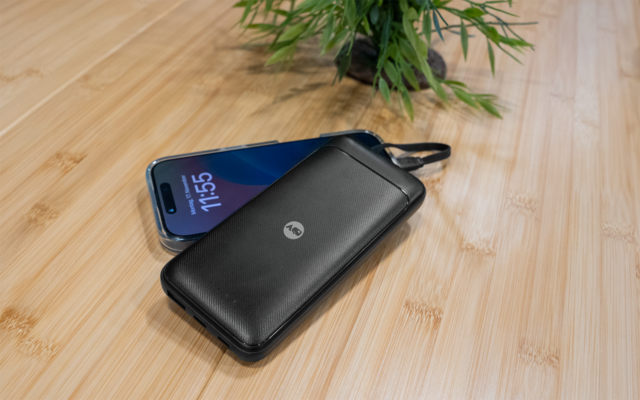 Batería externa negra con logo Motorola junto a un teléfono inteligente azul (pantalla 11:55) sobre una superficie de madera clara, planta verde al fondo