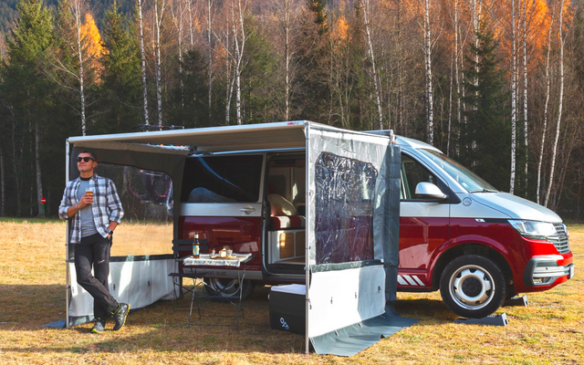 Rotes-weißes Wohnmobil mit aufgespanntem Vorzelt auf einer Wiese, eine Person lehnt daran, Klapptisch mit Tellern, Tassen und Flaschen, Birkenwald im Hintergrund
