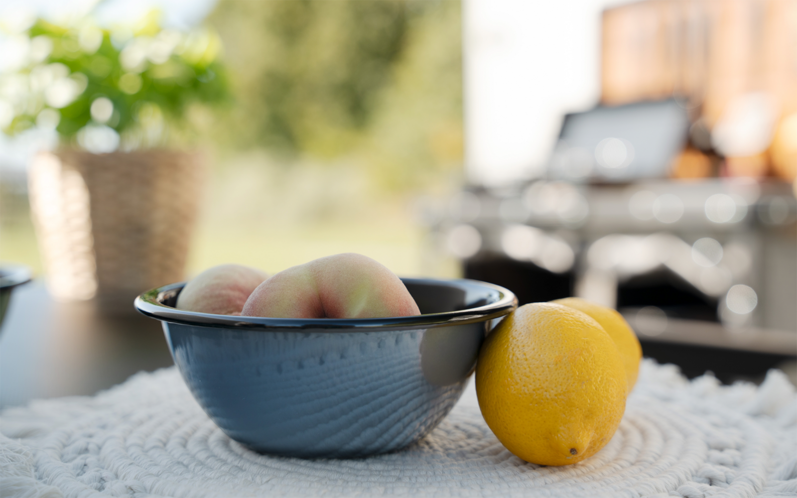 Bol bleu avec des pêches, un citron à côté sur un tissu blanc texturé ; arrière-plan flou avec un panier en osier et extérieur