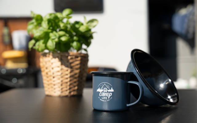 Blauer Emaille-Becher mit Aufschrift camp life und schräg gestellte schwarze Schüssel auf einem Tisch, links ein geflochtener Korb mit grüner Pflanze, unscharfer Hintergrund