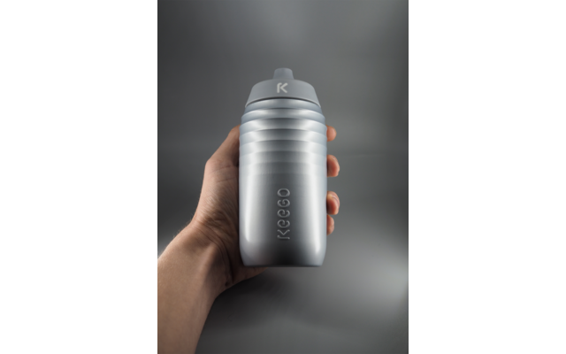 Hand houdt zilverkleurige drinkfles met horizontale ribbels en het reliëf "KEEGO", grijze schroefdop met "K"-logo, effen grijze achtergrond