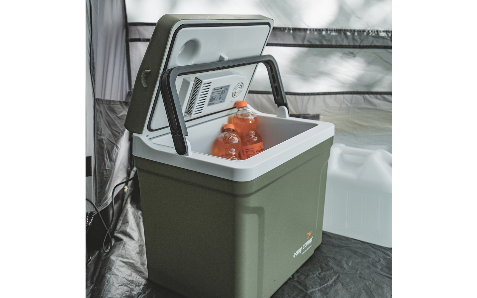 Glacière portable vert olive avec couvercle ouvert, poignée noire rabattable et intérieur blanc ; deux bouteilles de boisson orange à l'intérieur ; jerrican blanc et paroi en tissu gris en arrière-plan