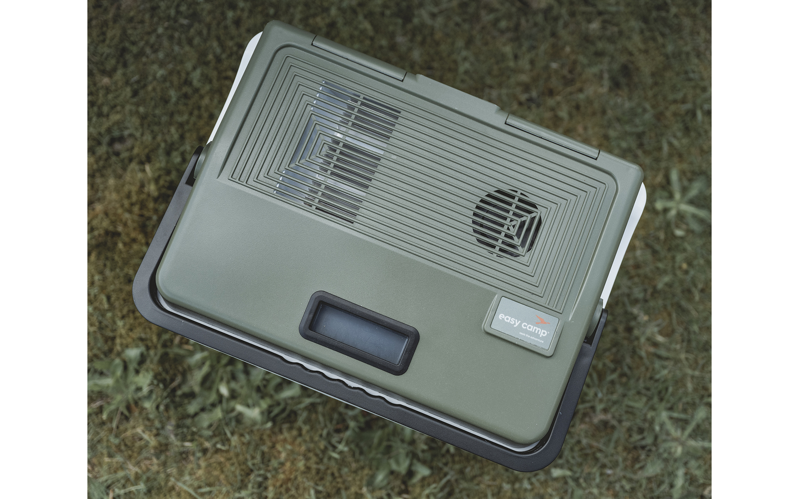 Boîtier rectangulaire en plastique vert-gris avec grilles de ventilation sur le dessus, petit écran rectangulaire, poignée noire à l'avant, autocollant, posé sur de l'herbe