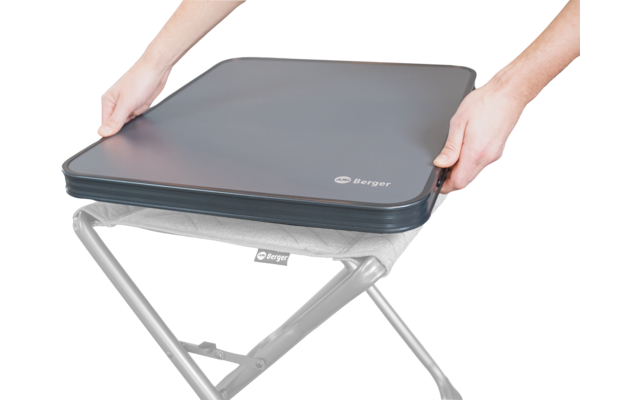 Two hands hold a gray padded square seat cover with a zipper and visible logo; below it a black crossed metal frame of a folding stool