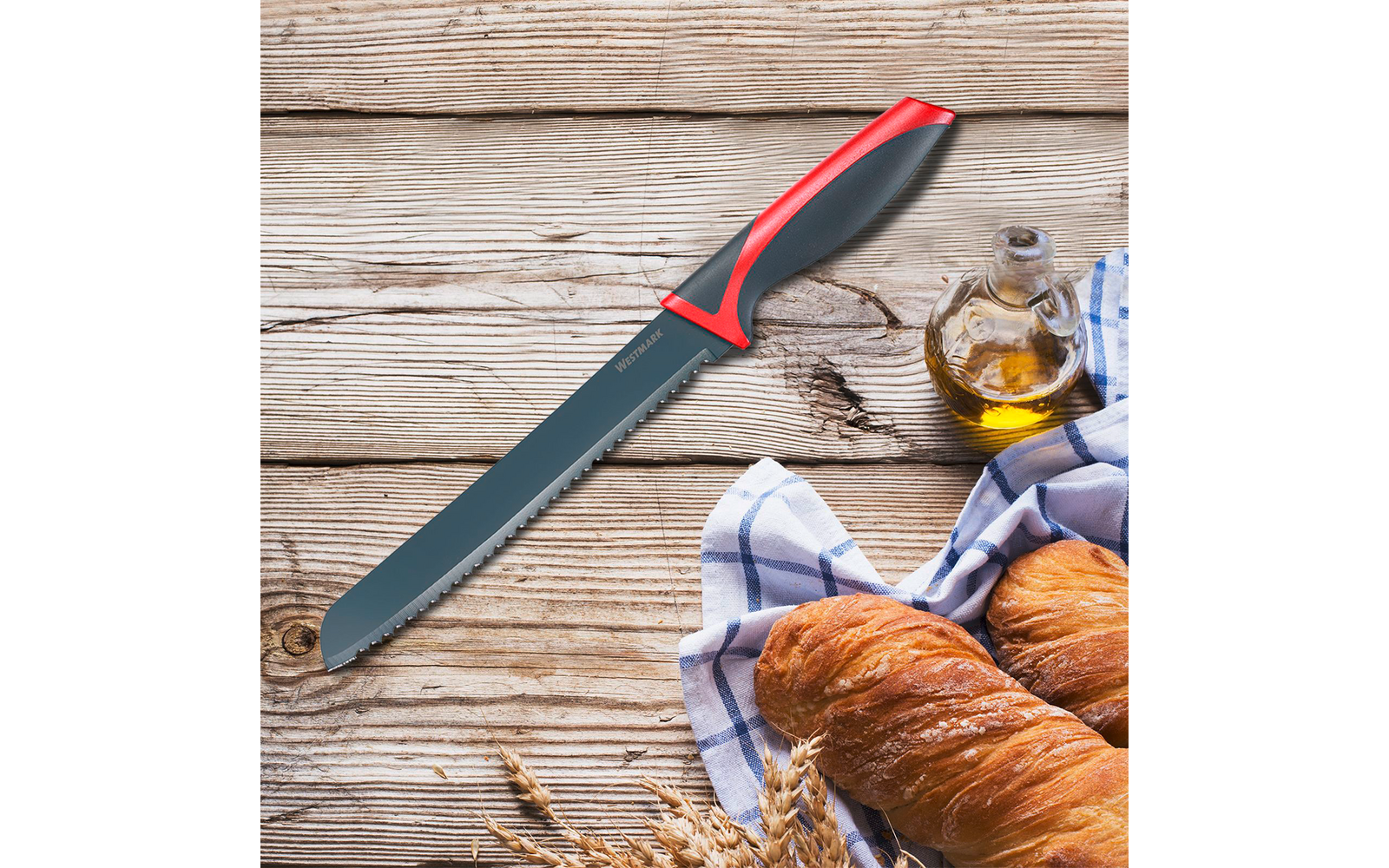 Auf einer Holzoberfläche liegt ein gezahntes Brotmesser mit rotem und schwarzem Griff, daneben zwei Baguettes in einem blau karierten Tuch, eine Glasflasche Olivenöl und einige Ähren
