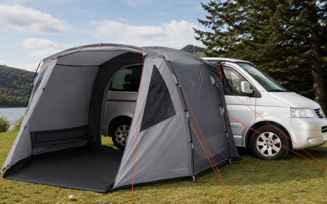 Furgone camper argentato su prato vicino a un lago, con veranda tenda grigia fissata e tiranti, alberi e colline sullo sfondo