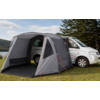 Furgone camper argentato su prato vicino a un lago, con veranda tenda grigia fissata e tiranti, alberi e colline sullo sfondo