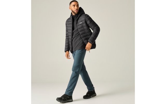 Persoon met zwarte donsjas en hoodie, petrolkleurige outdoorbroek en zwarte wandelschoenen, loopt voor een lichte achtergrond