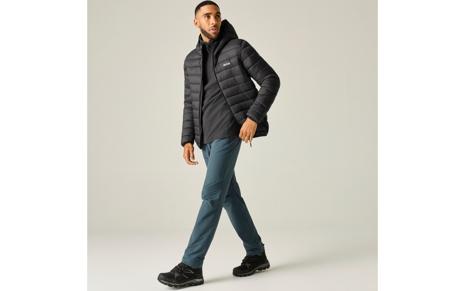 Persoon met zwarte donsjas en hoodie, petrolkleurige outdoorbroek en zwarte wandelschoenen, loopt voor een lichte achtergrond