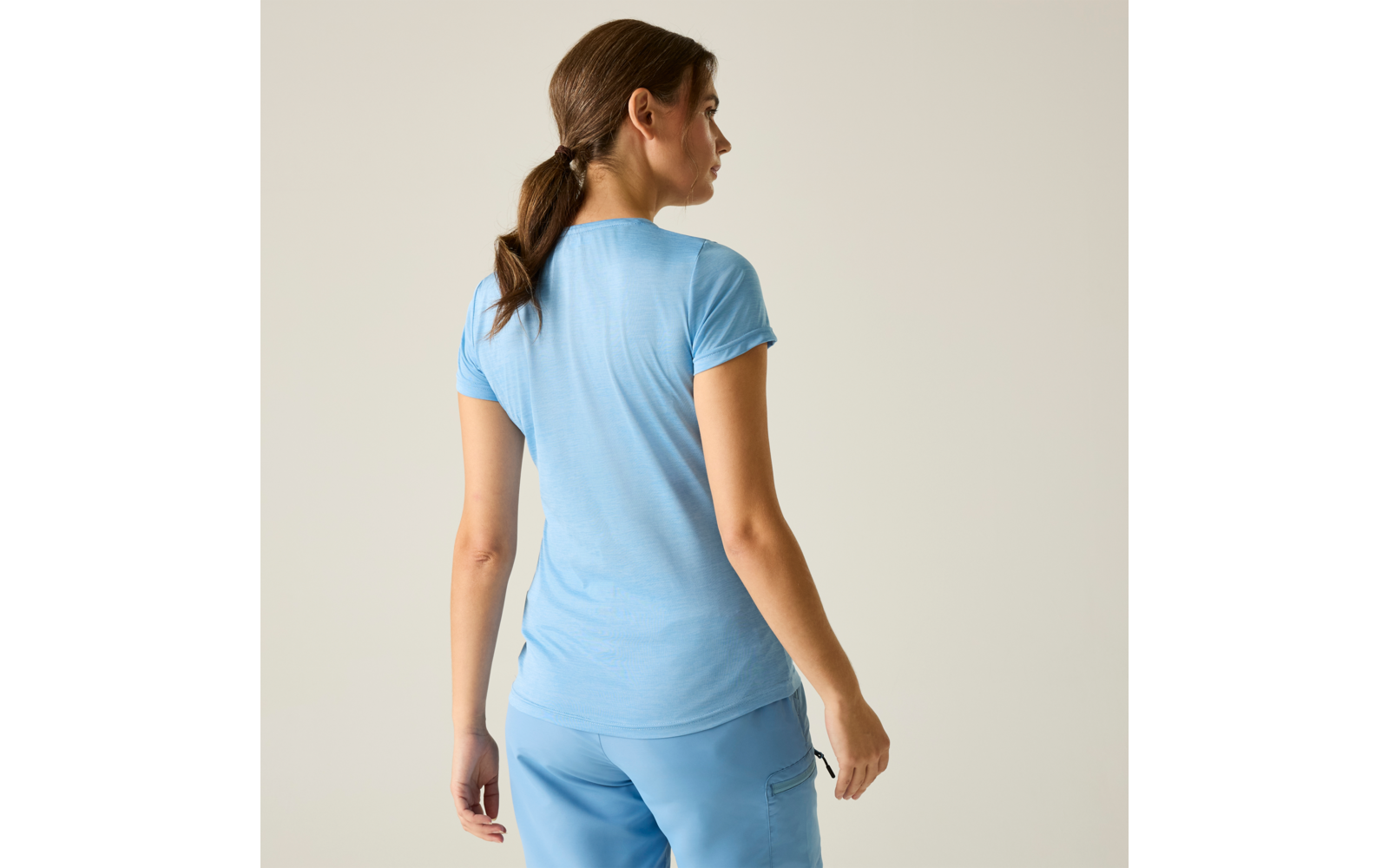 Persoon van achteren, draagt lichtblauw T-shirt met korte mouwen en lichtblauwe broek, haar in lage paardenstaart, effen lichte achtergrond