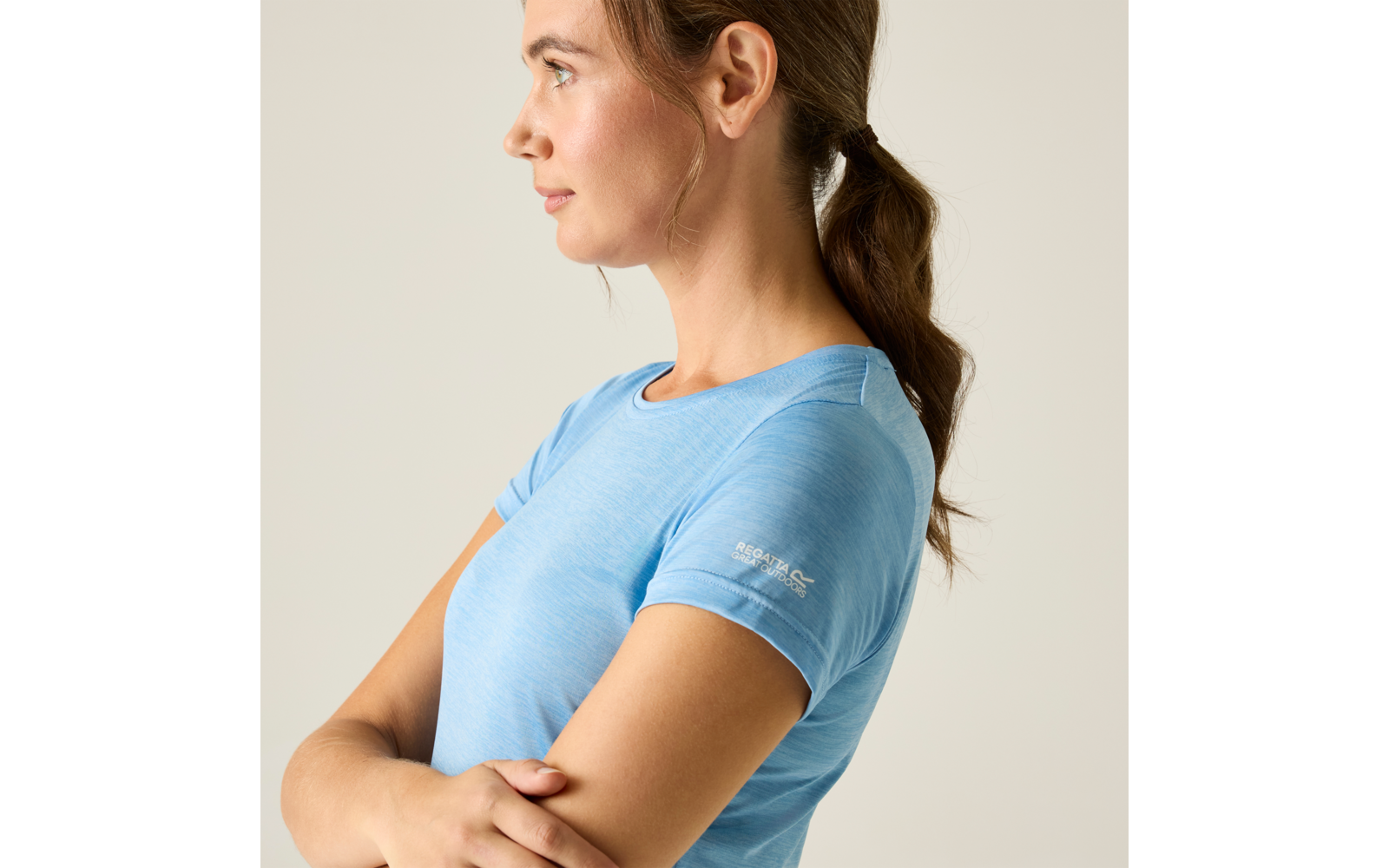 Zijaanzicht van een persoon die naar links kijkt met gekruiste armen, een lichtblauw T-shirt met korte mouwen en een lage paardenstaart voor een beige achtergrond