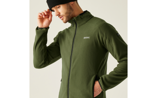 Ein Mann trägt eine grüne Jacke und eine schwarze Mütze. Er hält die Krempe der Mütze mit der Hand und schaut nach unten