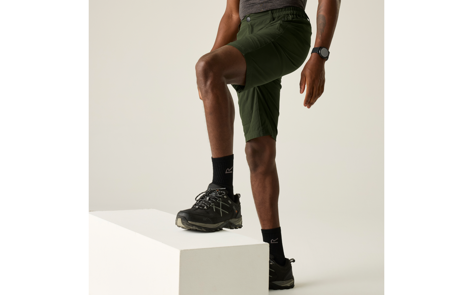 Person von der Taille abwärts, ein Fuß auf einem weißen Block; grüne Shorts, schwarze Socken mit Logo, schwarze Schuhe, Armbanduhr, heller neutraler Hintergrund