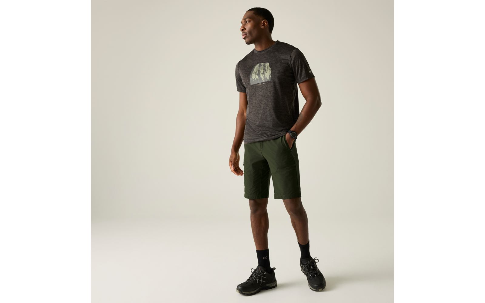 Ein stehender Mann in dunkelgrauem T-Shirt mit Motiv, grünen Shorts, schwarzen Socken und schwarzen Sneakers, mit Armbanduhr, Blick nach links vor hellem einfarbigem Hintergrund