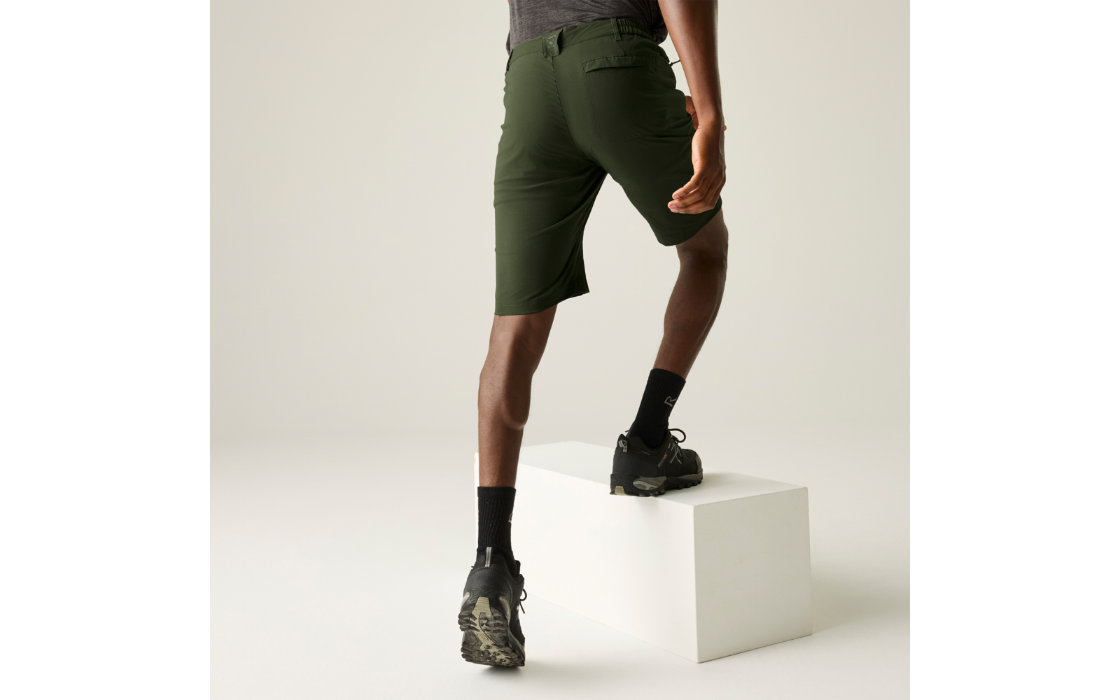 Unterkörper einer Person in olivgrünen knielangen Shorts, schwarzen Sportsocken und schwarzen Wanderschuhen; ein Fuß steht auf einem weißen rechteckigen Quader, neutraler Hintergrund, rechte Hand sichtbar