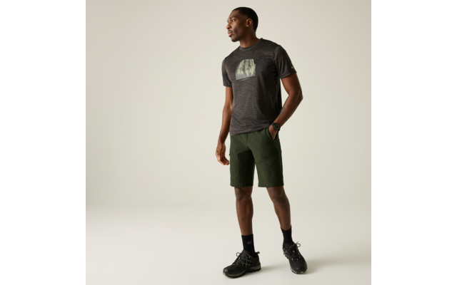 Ein stehender Mann in dunkelgrauem T-Shirt mit Motiv, grünen Shorts, schwarzen Socken und schwarzen Sneakers, mit Armbanduhr, Blick nach links vor hellem einfarbigem Hintergrund