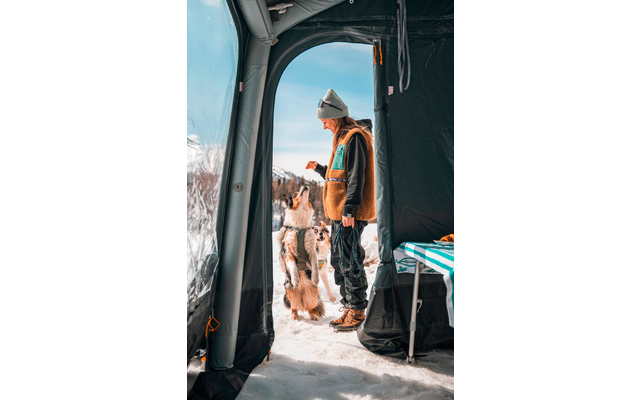 Persoon met muts en bruine jas staat bij de tentopening in de sneeuw en houdt een klein voorwerp voor een hond die op zijn achterpoten staat; tentinterieur en bergen zichtbaar