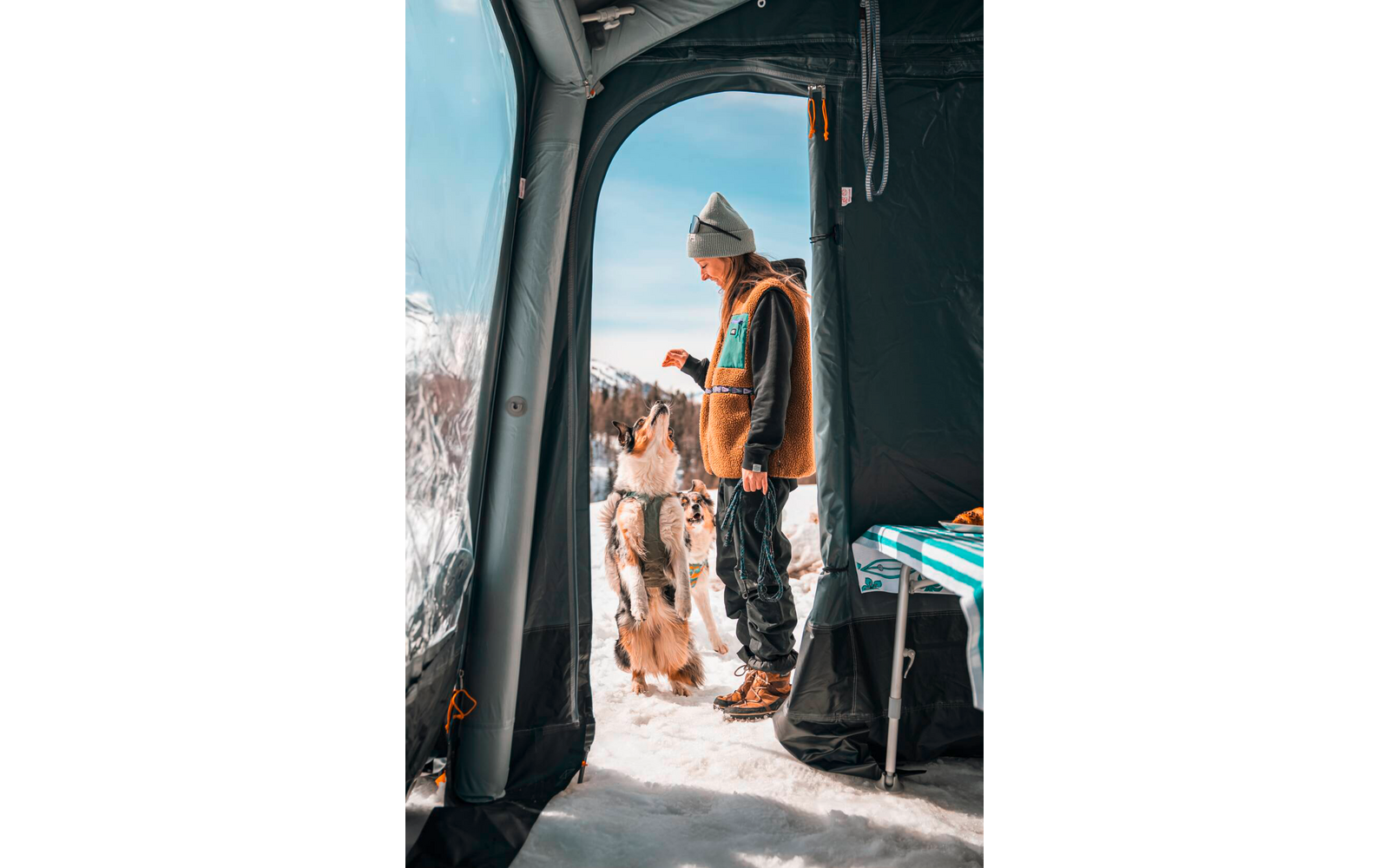 Persoon met muts en bruine jas staat bij de tentopening in de sneeuw en houdt een klein voorwerp voor een hond die op zijn achterpoten staat; tentinterieur en bergen zichtbaar