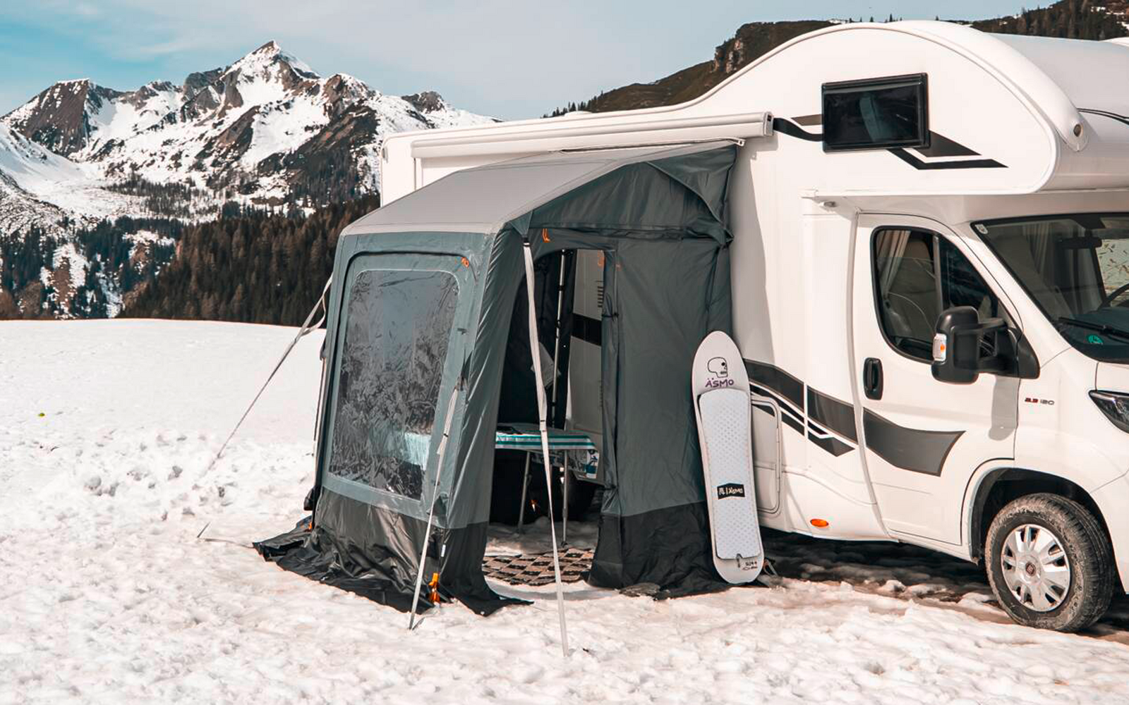 Witte camper met aangebouwde voortent op een met sneeuw bedekt veld, naaldbos en met sneeuw bedekte bergen op de achtergrond onder een blauwe lucht
