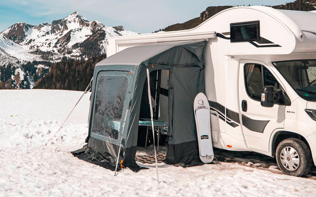 Witte camper met aangebouwde voortent op een met sneeuw bedekt veld, naaldbos en met sneeuw bedekte bergen op de achtergrond onder een blauwe lucht