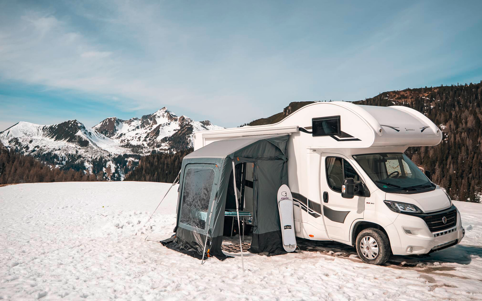White motorhome with attached awning on a snow-covered field, conifer forest and snow-capped mountains in the background under a blue sky
