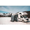 White motorhome with attached awning on a snow-covered field, conifer forest and snow-capped mountains in the background under a blue sky
