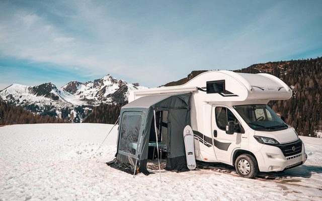 Weißes Wohnmobil mit angebautem Vorzelt auf schneebedecktem Feld, im Hintergrund Nadelwald und schneebedeckte Berge unter blauem Himmel